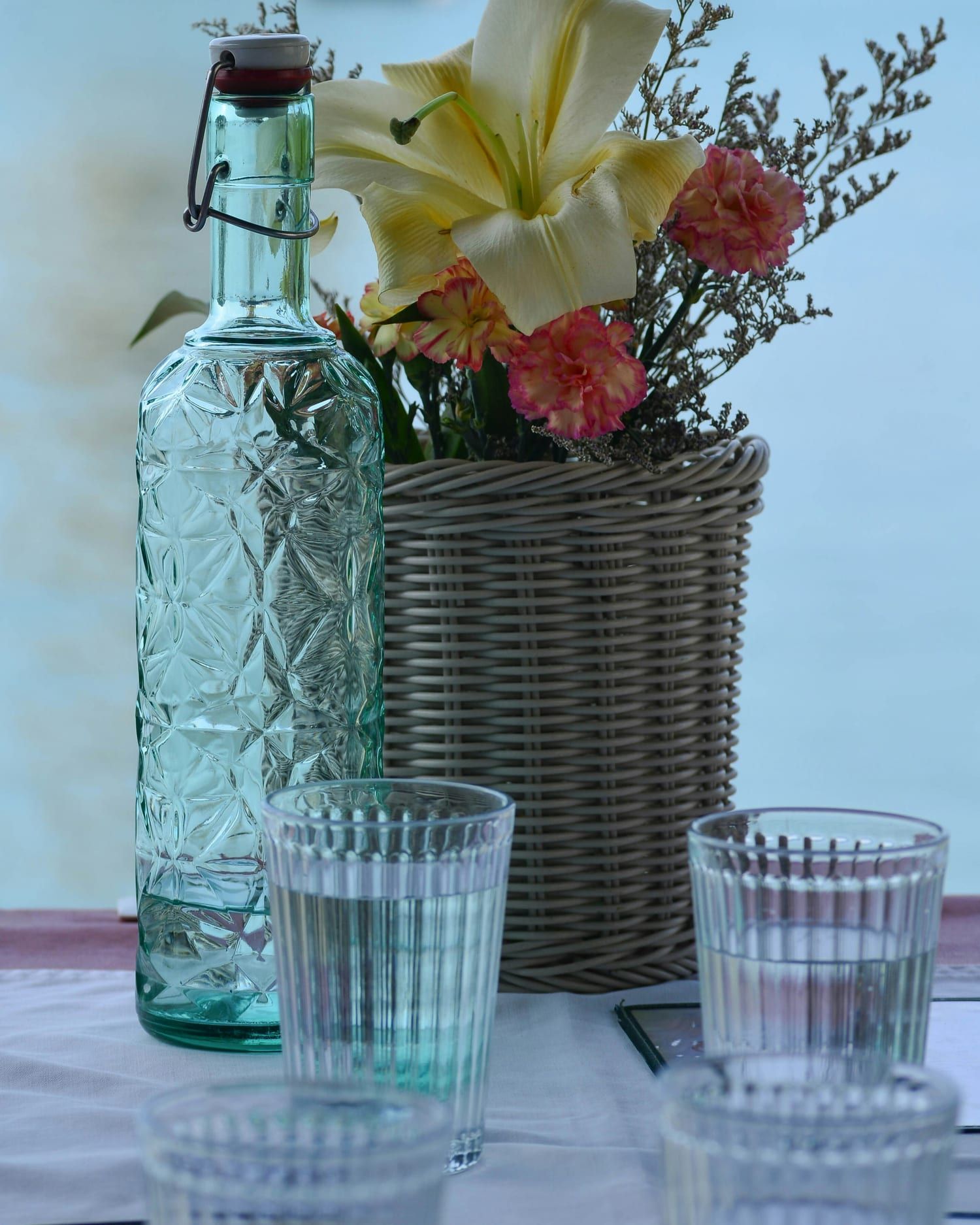 Bouteille de verre, lys et œillets dans un cache-pot tressé, verres d'eau sur une table, devant la mer